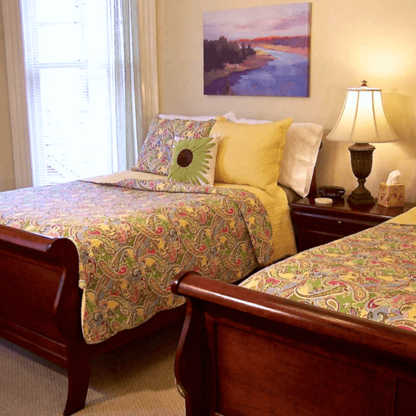 A cozy bedroom featuring two wooden beds with colorful paisley-patterned quilts, a lamp, and an abstract landscape painting on the wall.