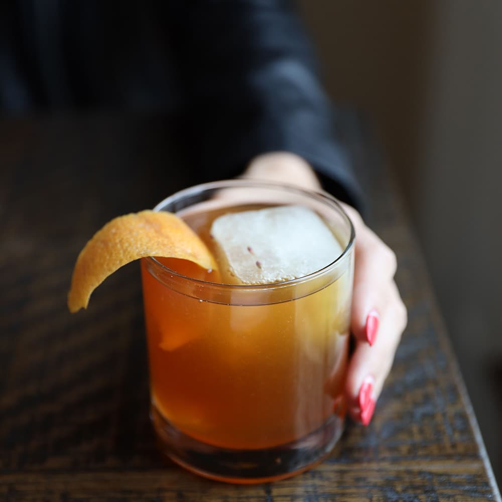 A hand with red nails holds a glass of cocktail garnished with an orange peel and an ice cube.