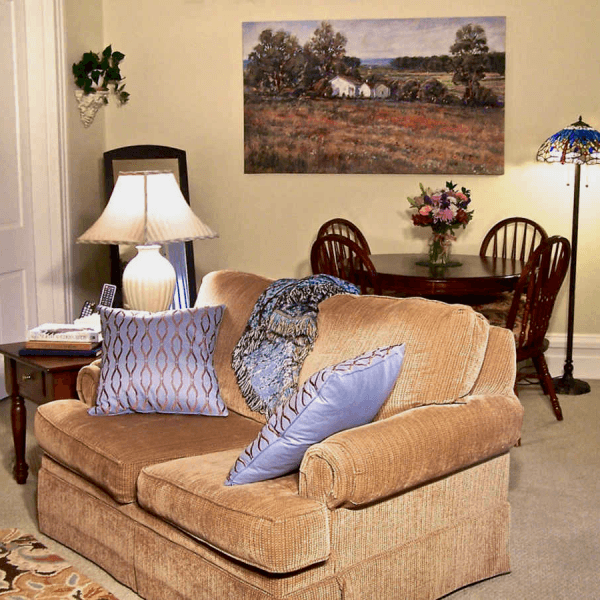 A cozy living room featuring a tan couch with decorative pillows, a lamp, a dining table with flowers, and a landscape painting on the wall.