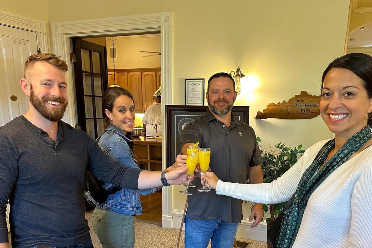 A group of four people clink glasses filled with mimosas, smiling in a warmly lit room.