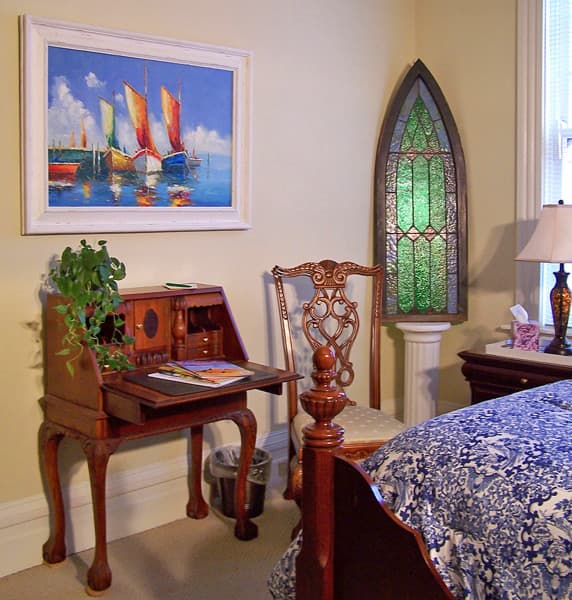 A cozy bedroom featuring a wooden desk, an ornate chair, a vibrant painting of boats, and a stained glass window.