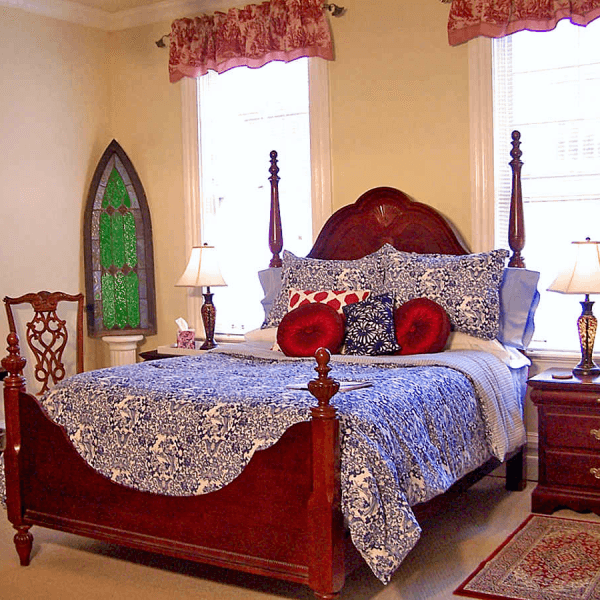 A cozy bedroom featuring a four-poster bed with blue and white bedding, decorative pillows, and a stained glass window.