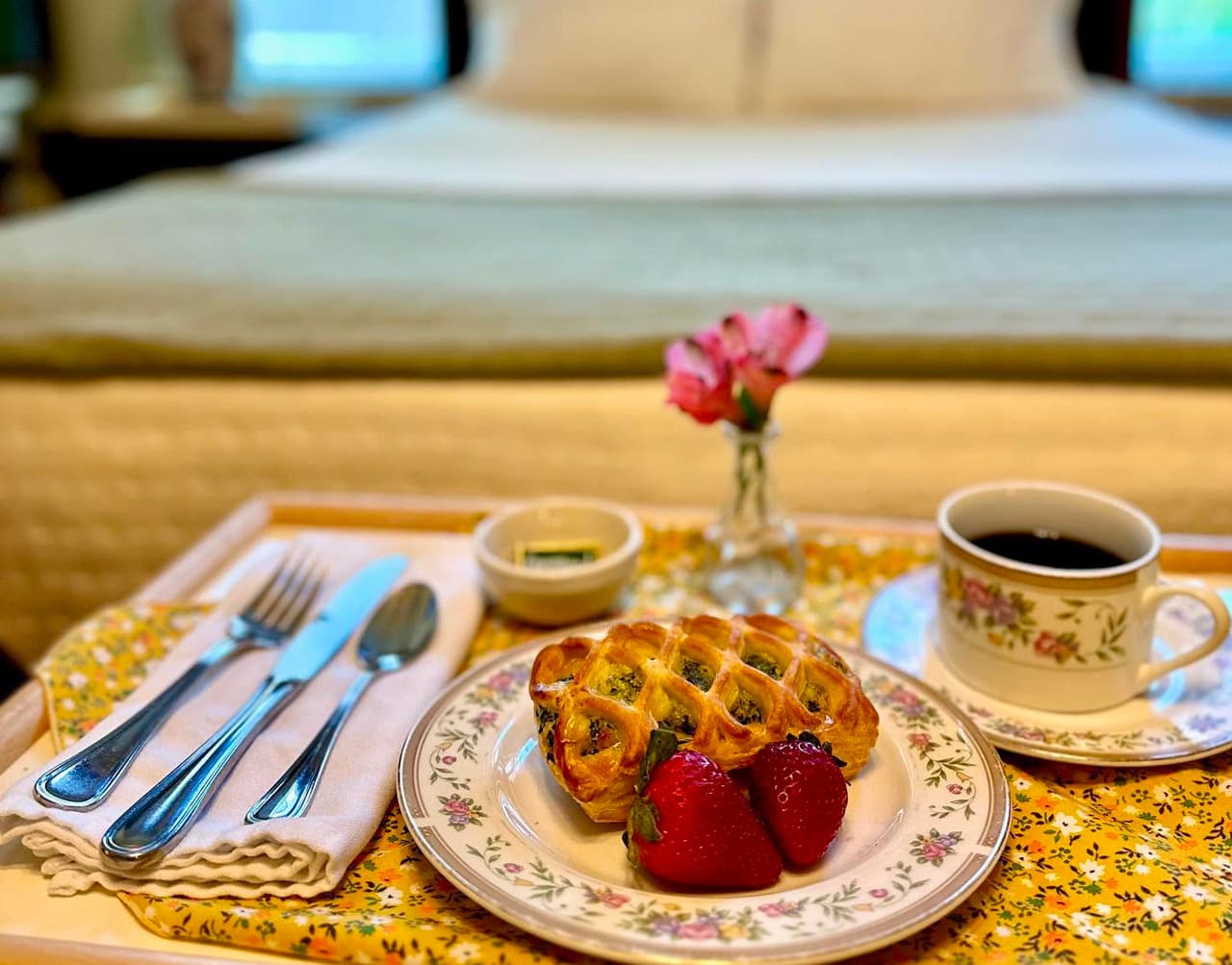 A breakfast tray with a pastry, strawberries, coffee, and a flower arrangement sits in front of a neatly made bed.