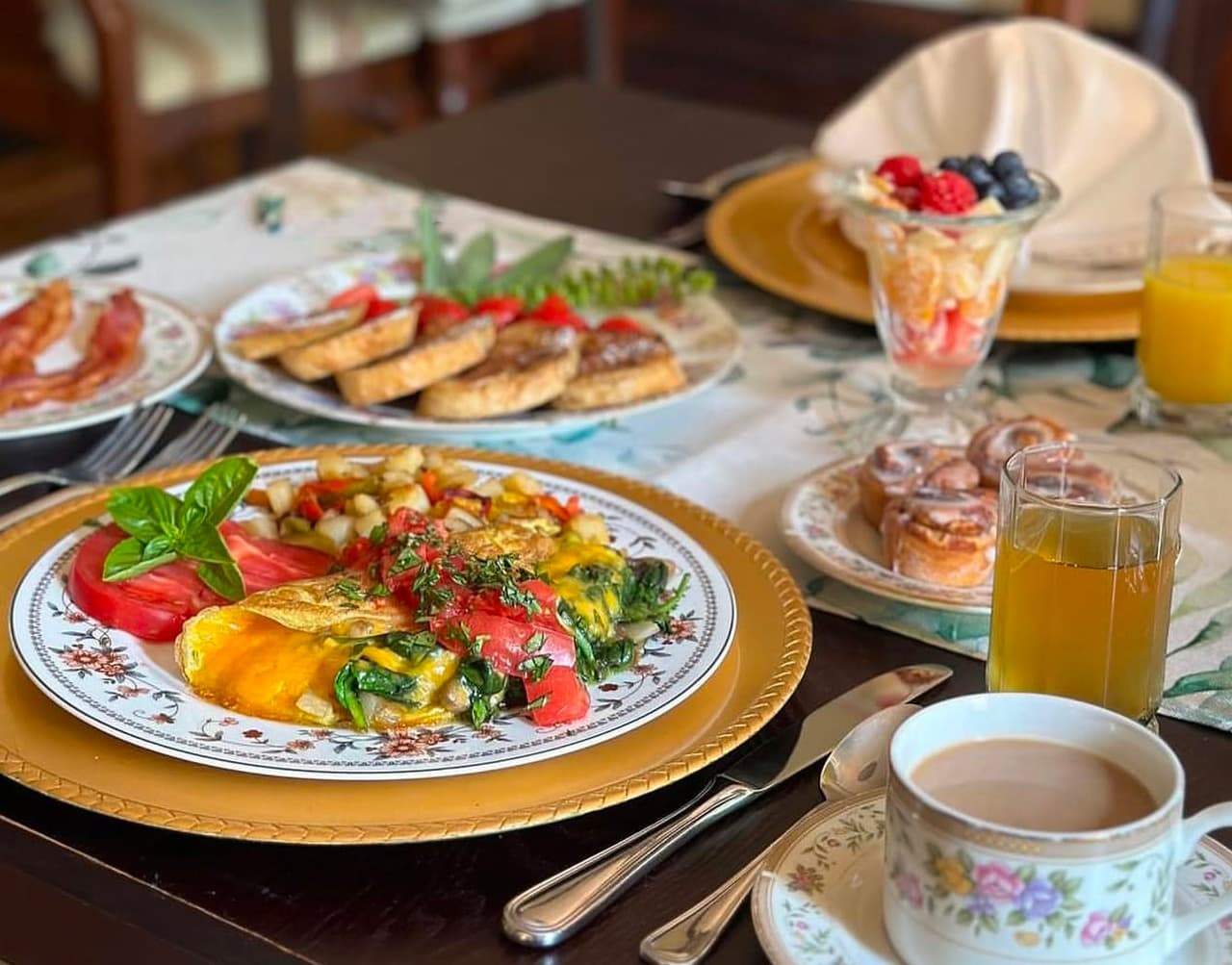 A beautifully arranged breakfast table featuring an omelette, diced potatoes, bacon, cookies, a fruit parfait, cinnamon rolls, orange juice, and coffee.
