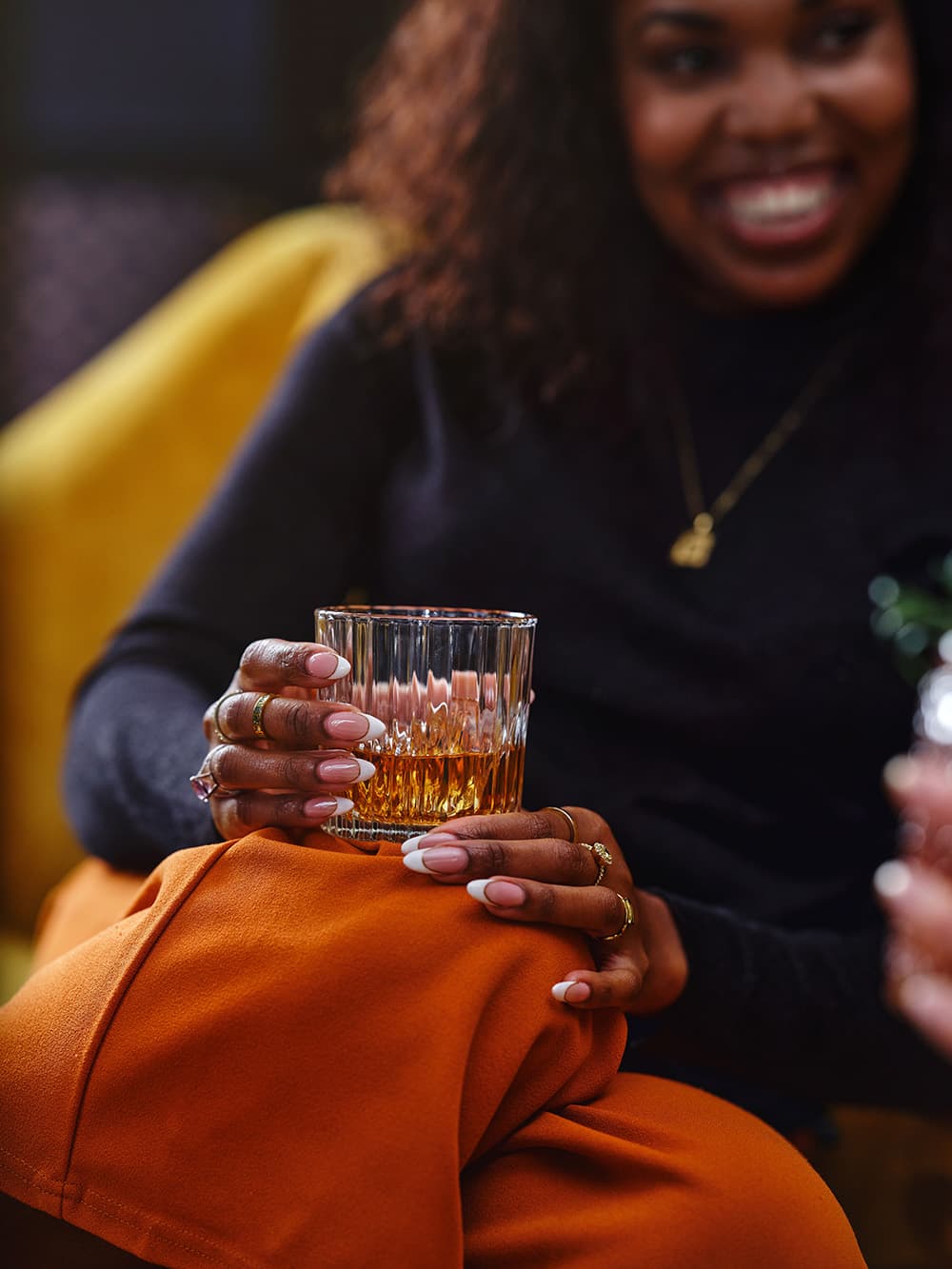 A woman casually holds a glass of whiskey while sitting on a couch, wearing an orange outfit and smiling.