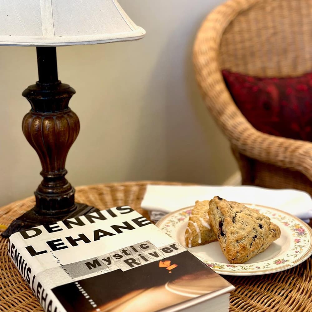 A table with a lamp, a book titled "Mystic River" by Dennis Lehane, and a plate of scones.