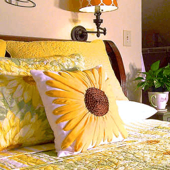 A cozy bed adorned with yellow floral pillows and a bright sunflower cushion, beside a potted plant.