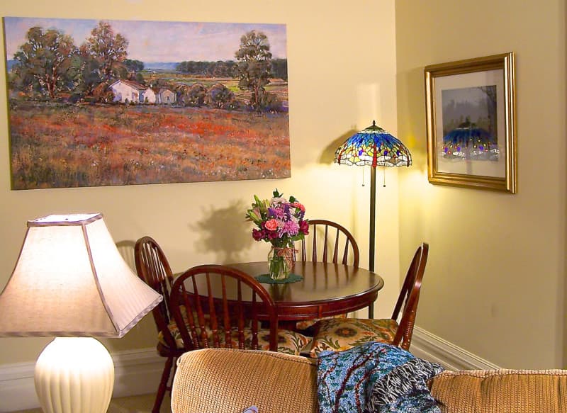 A cozy corner dining area featuring a floral painting, a vase of flowers on the table, and a stained glass lamp.