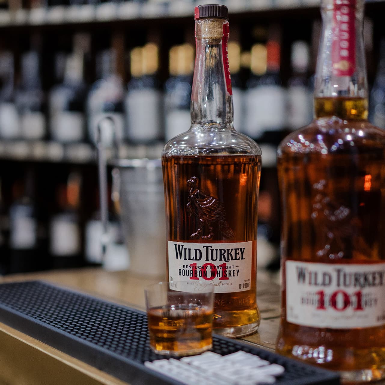 Bottles of Wild Turkey 101 bourbon whiskey are displayed on a bar with a glass of whiskey in front.