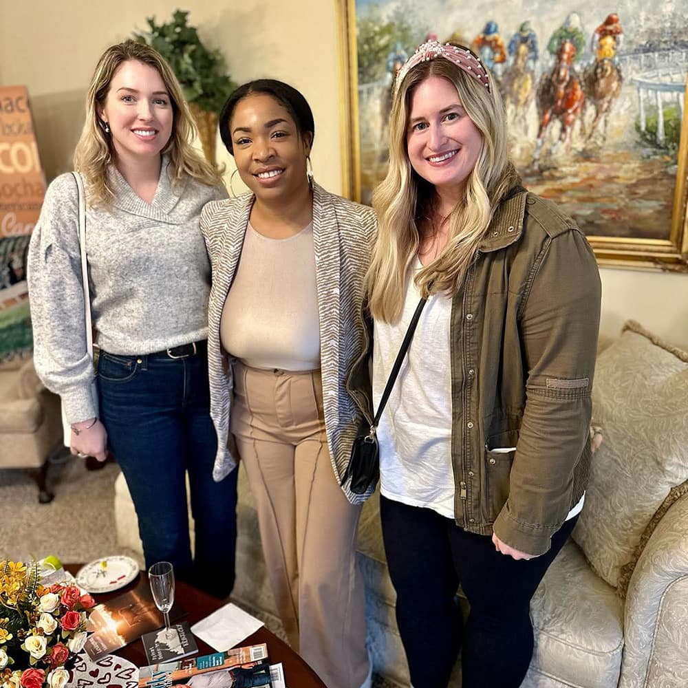 Three women stand together, smiling in a warmly decorated room with a painting in the background.