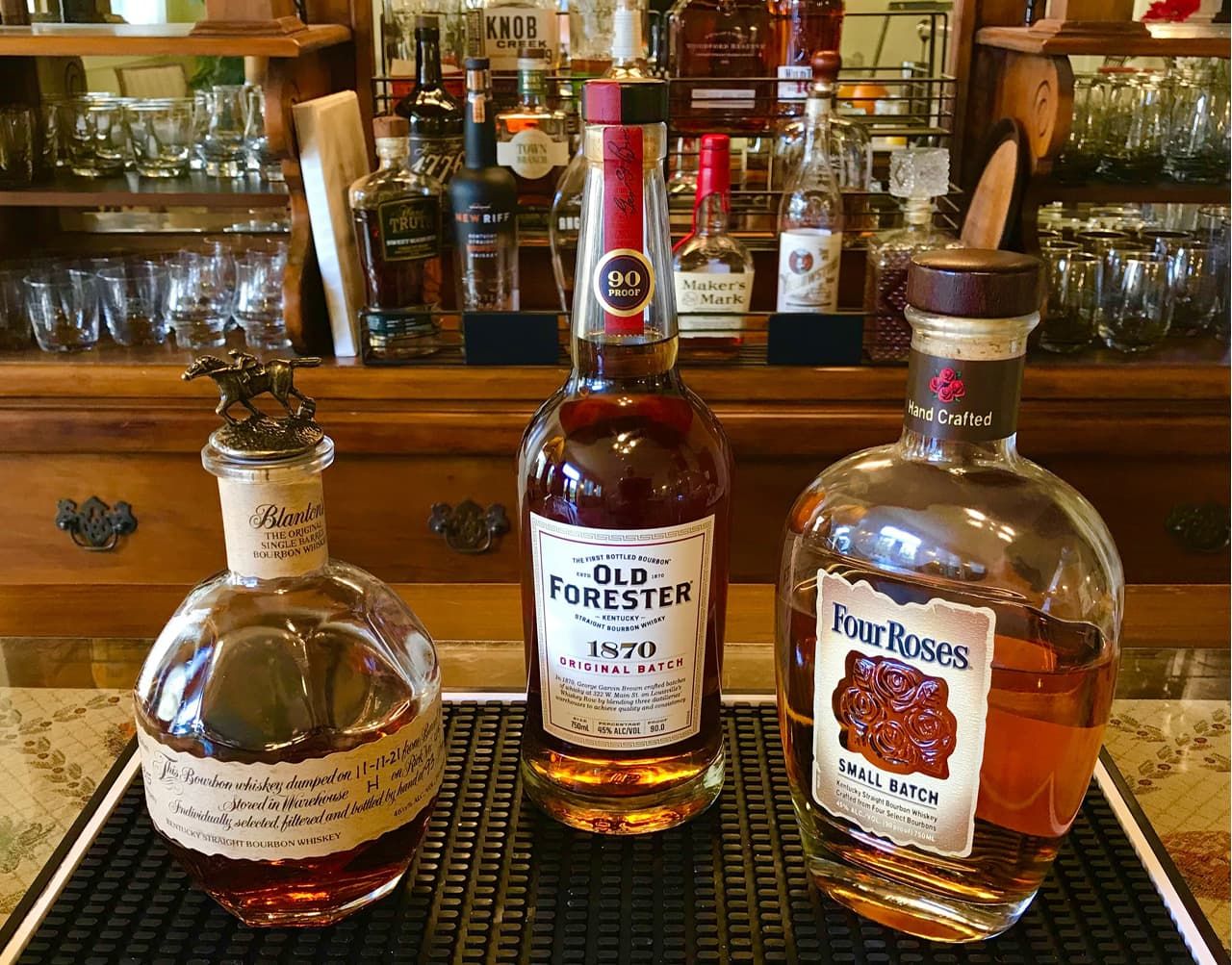 Three bourbon bottles displayed on a bar with various glasses in the background.
