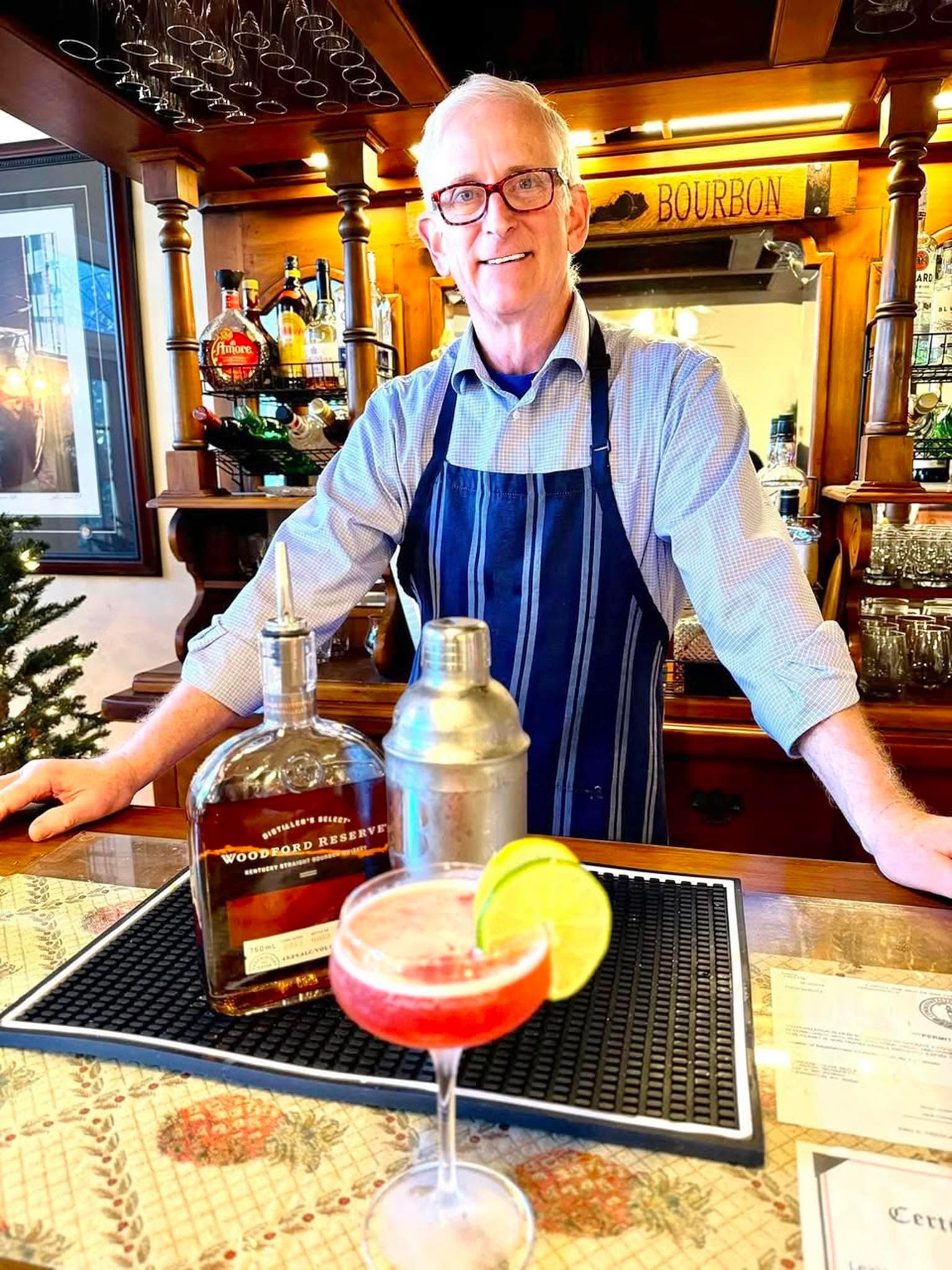 A smiling bartender in an apron stands behind a bar with a cocktail and a bottle of Woodford Reserve bourbon.
