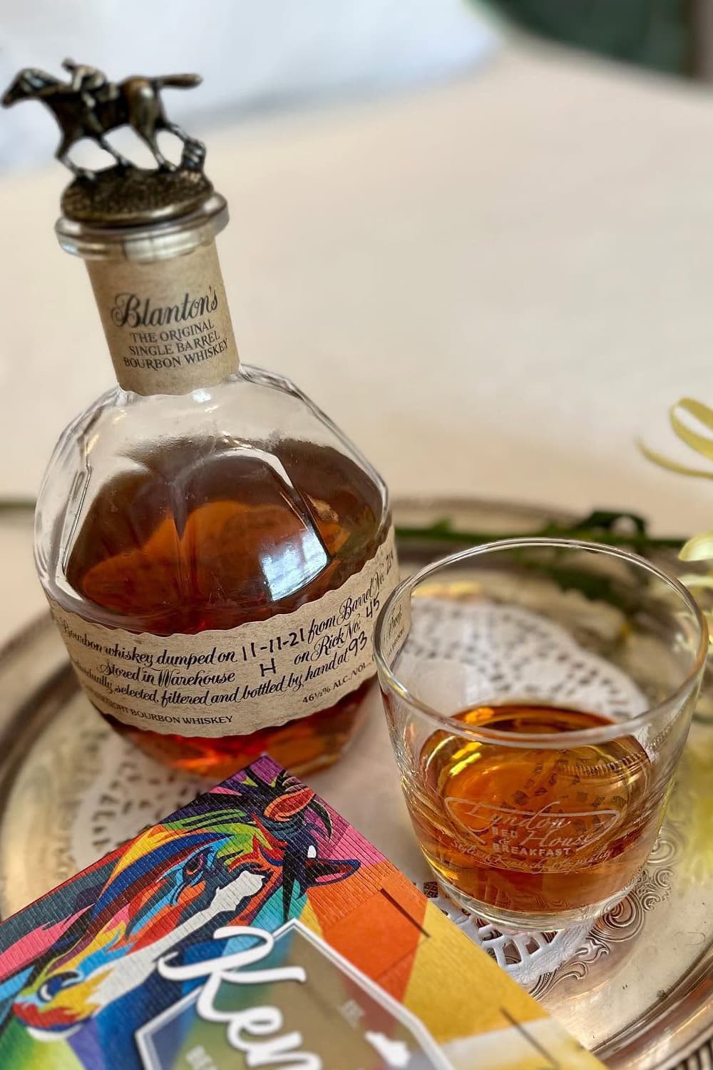 A bottle of Blanton's bourbon whiskey with a glass of whiskey and a colorful book on a decorative tray.
