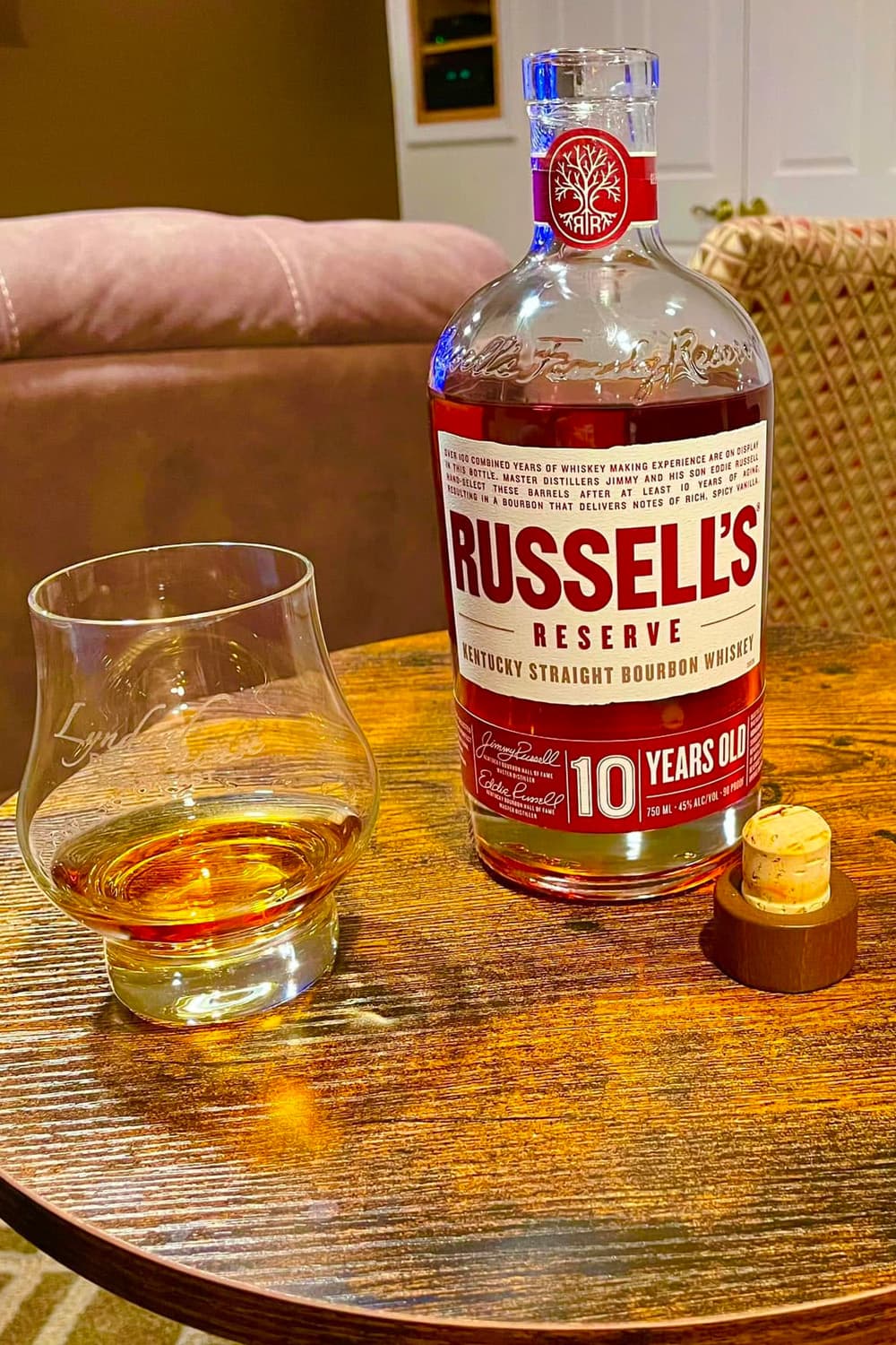 A bottle of Russell's Reserve bourbon sits next to a glass with a pour on a wooden table.