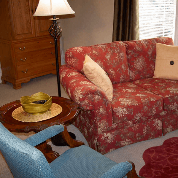 A cozy living room scene featuring a floral-patterned red couch, a blue armchair, a wooden side table with a decorative bowl, and a lamp.