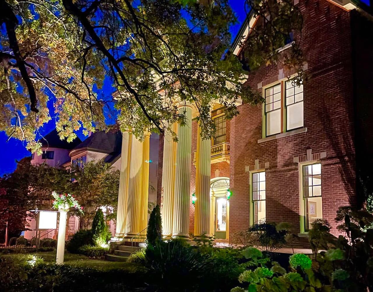 A historic brick building illuminated at night, featuring tall columns and lush greenery.