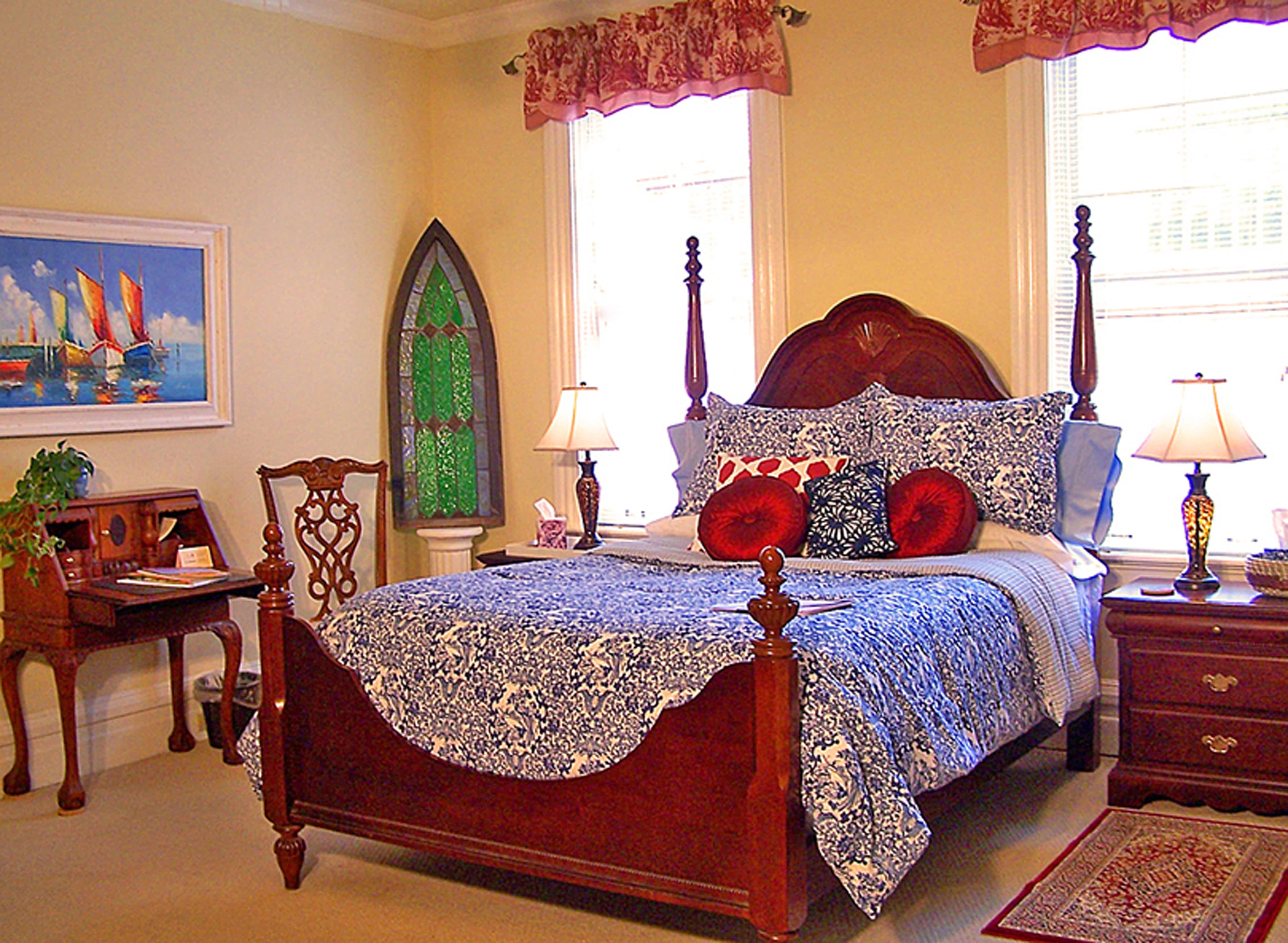 A cozy bedroom featuring a wooden bed with blue floral bedding, decorative pillows, a small desk, and natural light from large windows.