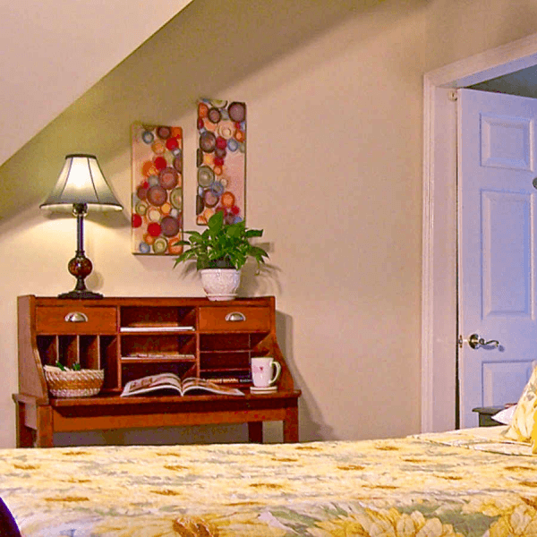 A cozy bedroom featuring a floral bedspread, wooden desk with a lamp, a potted plant, and colorful wall decor.