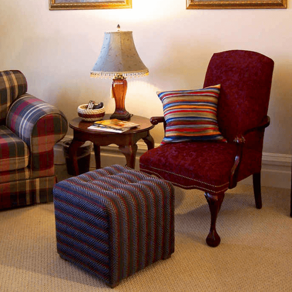 A cozy living room corner featuring a red armchair with a striped pillow, a patterned ottoman, a lamp, and a wooden side table.