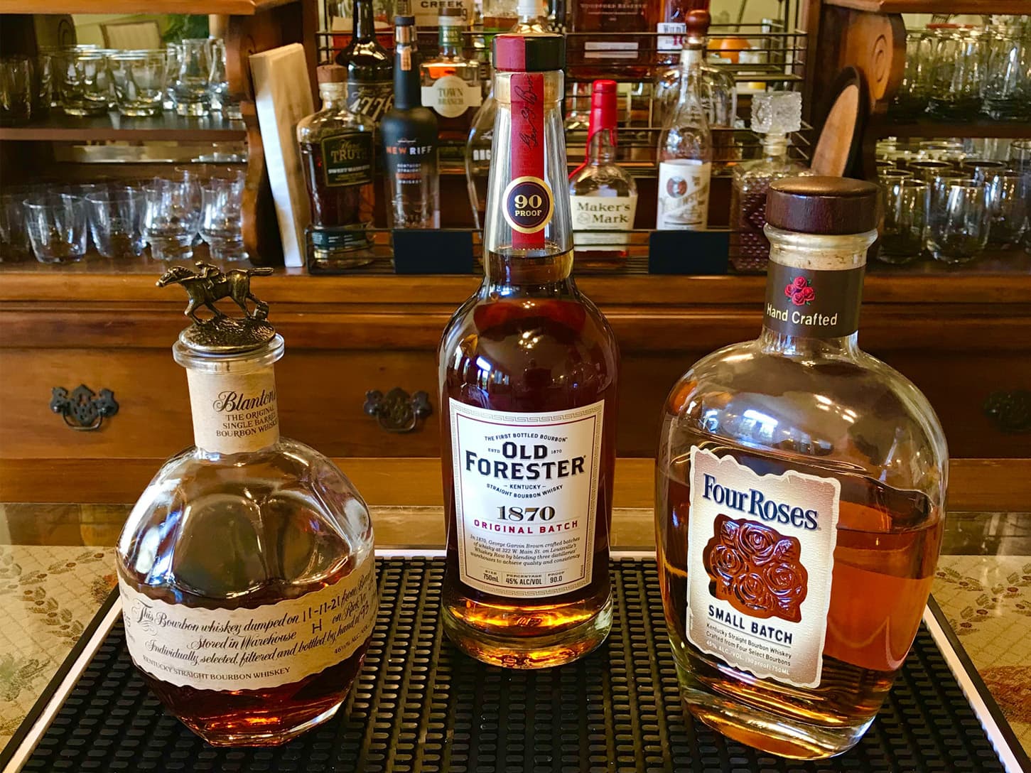 Three whiskey bottles-Blanton's, Old Forester 1870, and Four Roses Small Batch-on a black mat in front of a wooden bar cabinet filled with glassware.