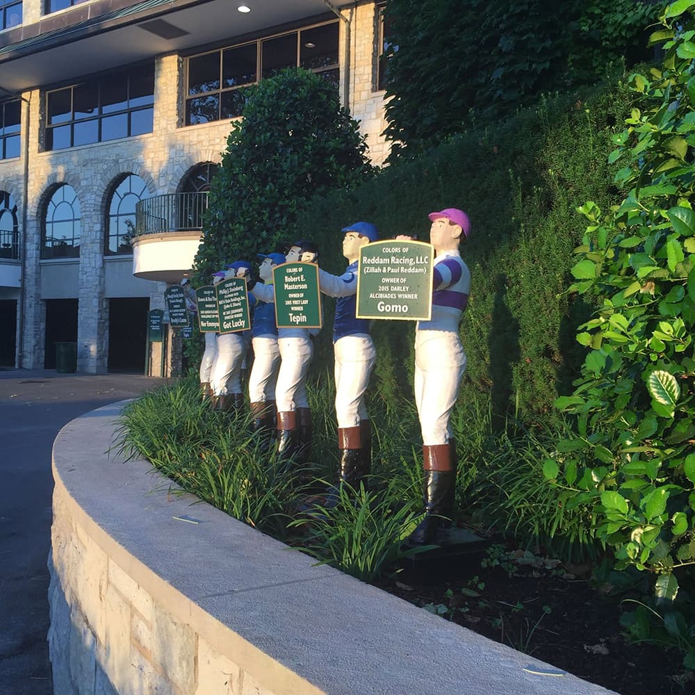 Line of jockey statues holding signs in front of a building, surrounded by greenery.