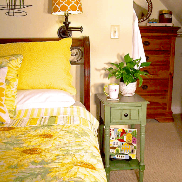 A cozy bedroom corner featuring a bed with bright yellow pillows, a small green nightstand, a potted plant, and a decorative mug.