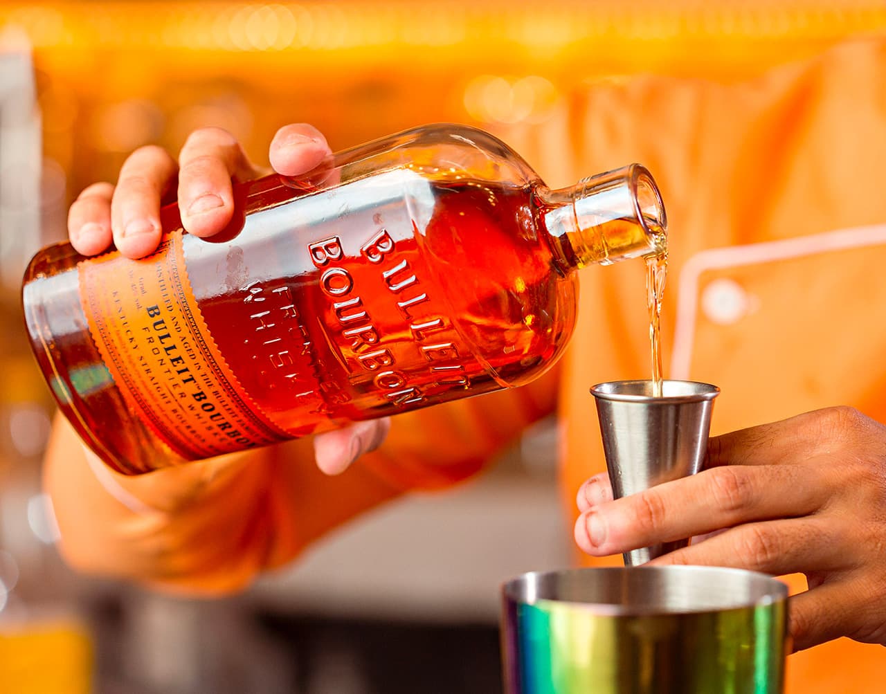 A bartender pours bourbon from a bottle into a jigger.