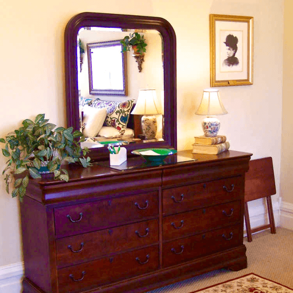 A wooden dresser with a mirror, two lamps, a potted plant, and a few books against a softly decorated wall.