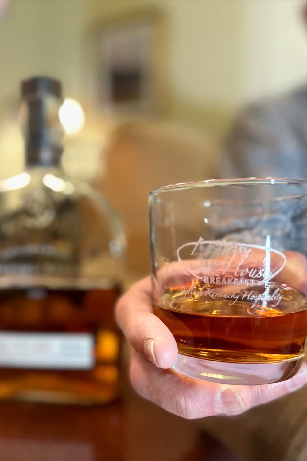 A person holds a glass of whiskey in front of a bottle.