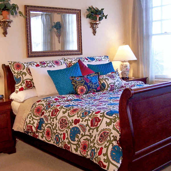 A cozy bedroom featuring a wooden sleigh bed with a colorful floral comforter and decorative pillows, accompanied by a bedside lamp and mirror.