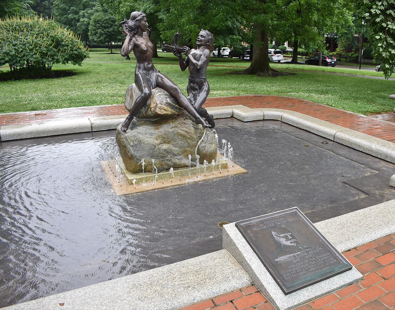 A bronze sculpture of two figures playing music, set on a rock in a fountain surrounded by greenery.