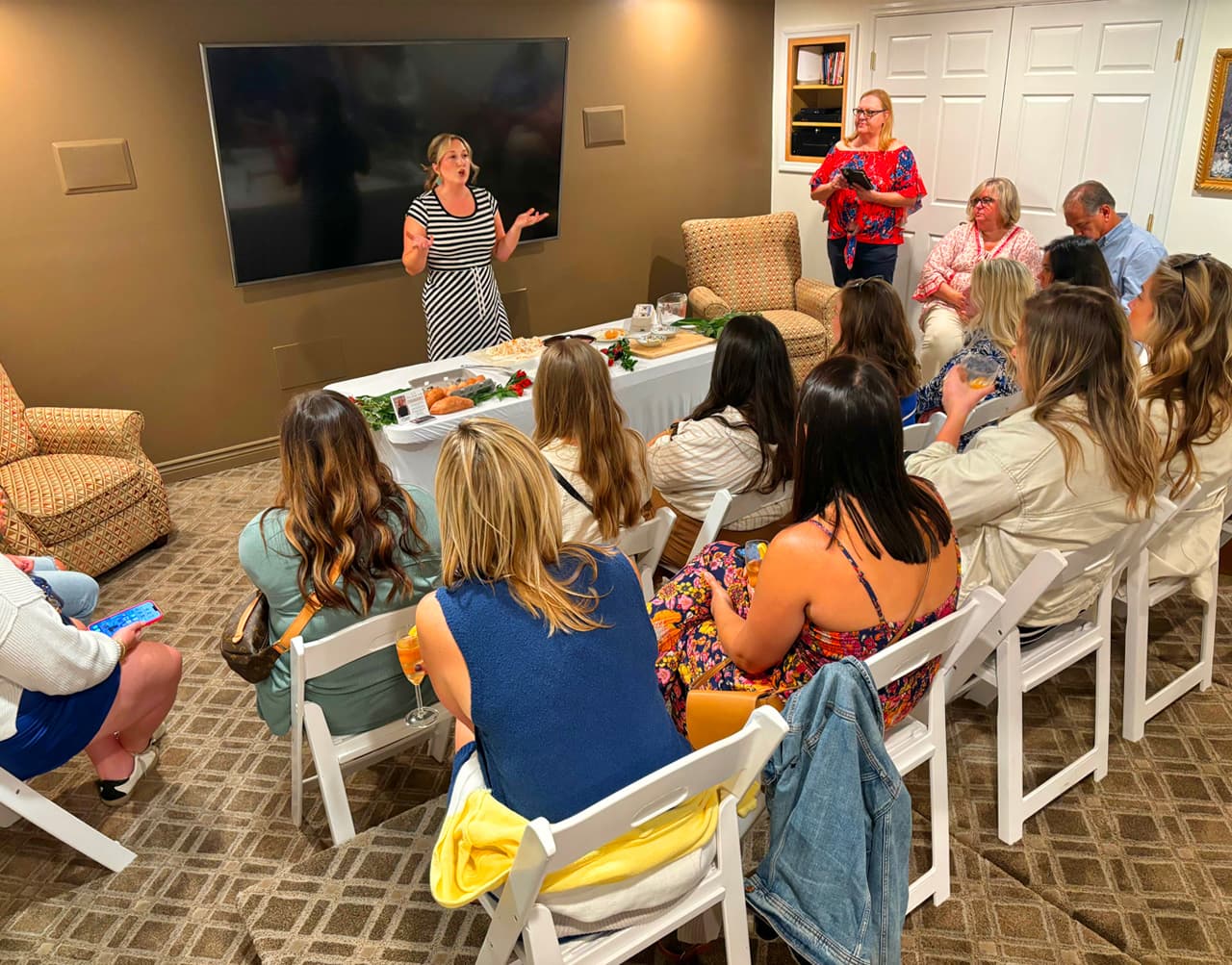 A woman in a striped dress speaks to an audience seated in a cozy room.