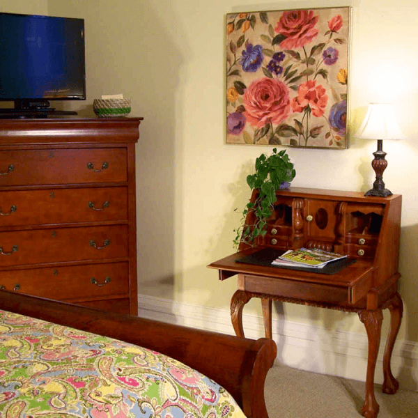 A cozy bedroom featuring a wooden dresser, a floral painting, a small writing desk, and a vibrant bedspread.